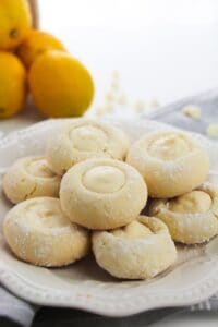 A plate of powdered sugar-dusted Lemon White Chocolate Cookies filled with creamy lemon filling, with whole lemons in the background.