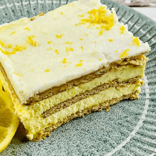 A slice of Lemon Icebox Cake on a green plate, garnished with lemon zest and a lemon slice. The cake has visible layers of crackers and lemon filling.