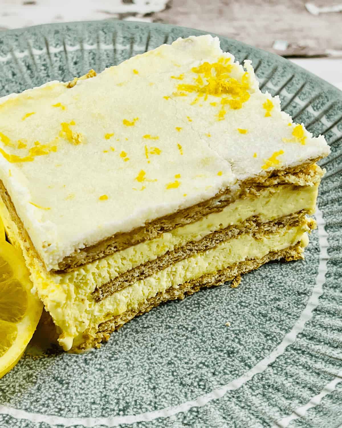 A slice of Lemon Icebox Cake on a green plate, garnished with lemon zest and a lemon slice. The cake has visible layers of crackers and lemon filling.