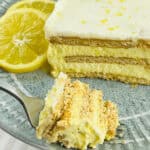 A slice of layered Lemon Icebox Cake with cream and graham crackers on a plate, garnished with two lemon slices, with a fork holding a piece in the foreground.