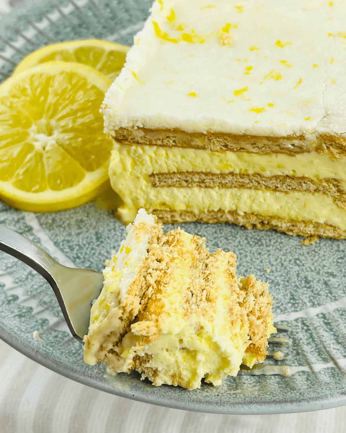 A slice of layered Lemon Icebox Cake with cream and graham crackers on a plate, garnished with two lemon slices, with a fork holding a piece in the foreground.