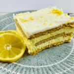 A slice of lemon cheesecake with layers of cream and crust is served on a green plate, reminiscent of a Lemon Icebox Cake, accompanied by a lemon slice garnish.