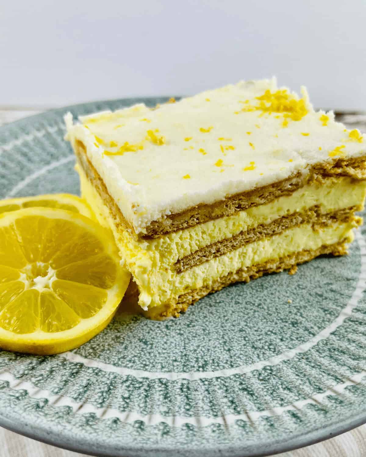 A slice of lemon cheesecake with layers of cream and crust is served on a green plate, reminiscent of a Lemon Icebox Cake, accompanied by a lemon slice garnish.