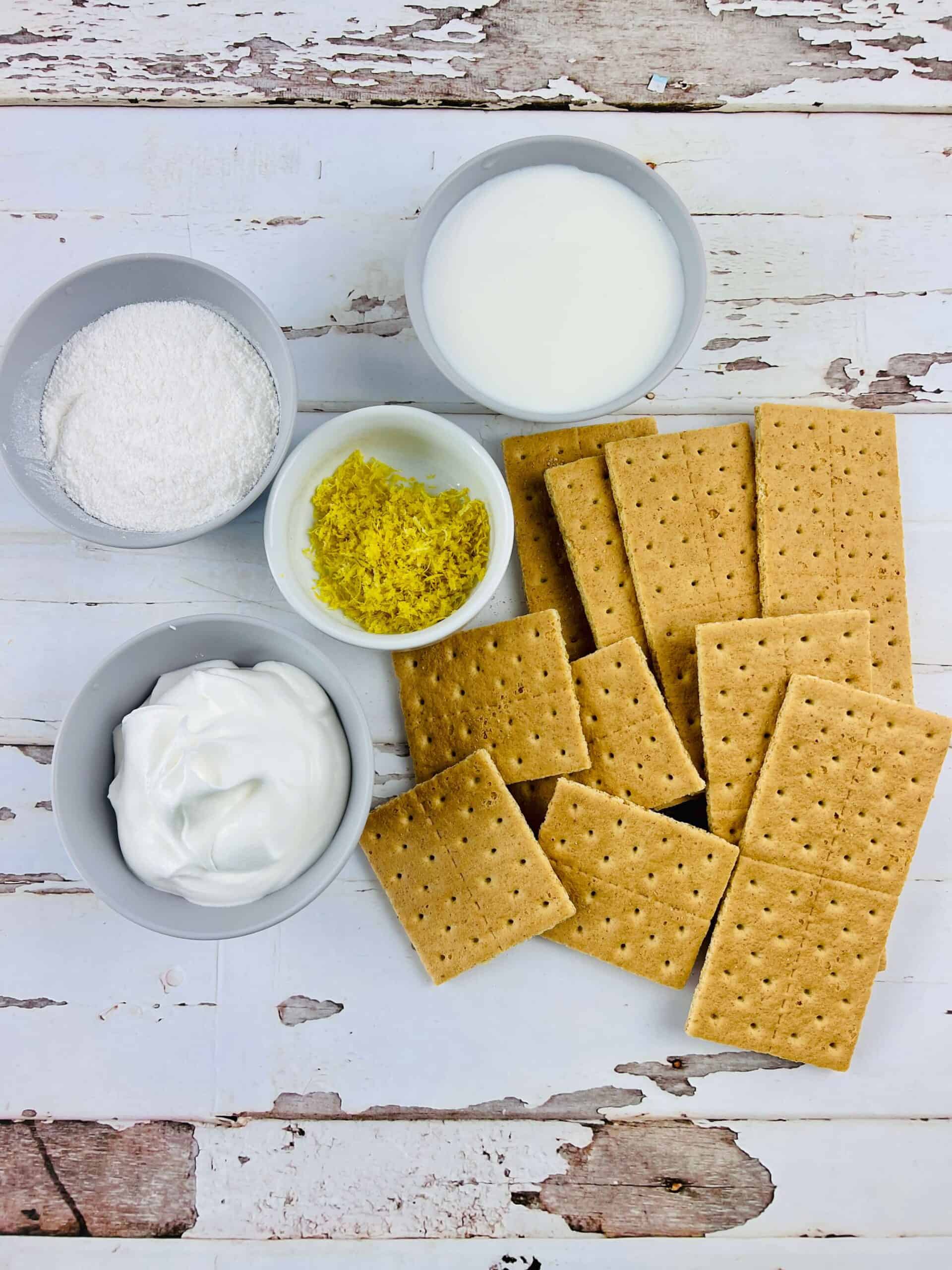 Ingredients for a Lemon Icebox Cake include graham crackers, whipped topping, powdered sugar, milk, and lemon zest, neatly arranged in bowls on a wooden surface.