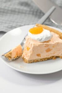 A slice of creamy Orange Cream Pie topped with a dollop of whipped cream and a small orange segment, served on a white plate with a fork.