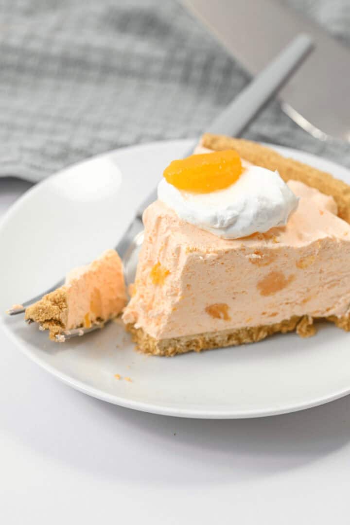 A slice of creamy Orange Cream Pie topped with a dollop of whipped cream and a small orange segment, served on a white plate with a fork.