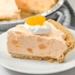 A slice of creamy dessert with chunks of fruit is on a white plate. The pie is topped with a dollop of whipped cream and an orange slice. A larger pie in a metal dish is in the background.