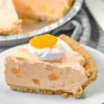 A slice of peach pie with a graham cracker crust, topped with a dollop of whipped cream and a peach chunk, reminiscent of an Orange Cream Pie, served on a white plate.