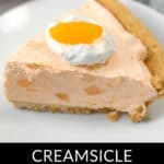 A slice of no-bake Orange Cream Pie with a graham cracker crust, topped with whipped cream and an orange segment, sits on a white plate.