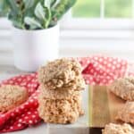 A stack sits on a white and wooden board, with more cookies scattered around. A red polka dot cloth and a potted plant are in the background.