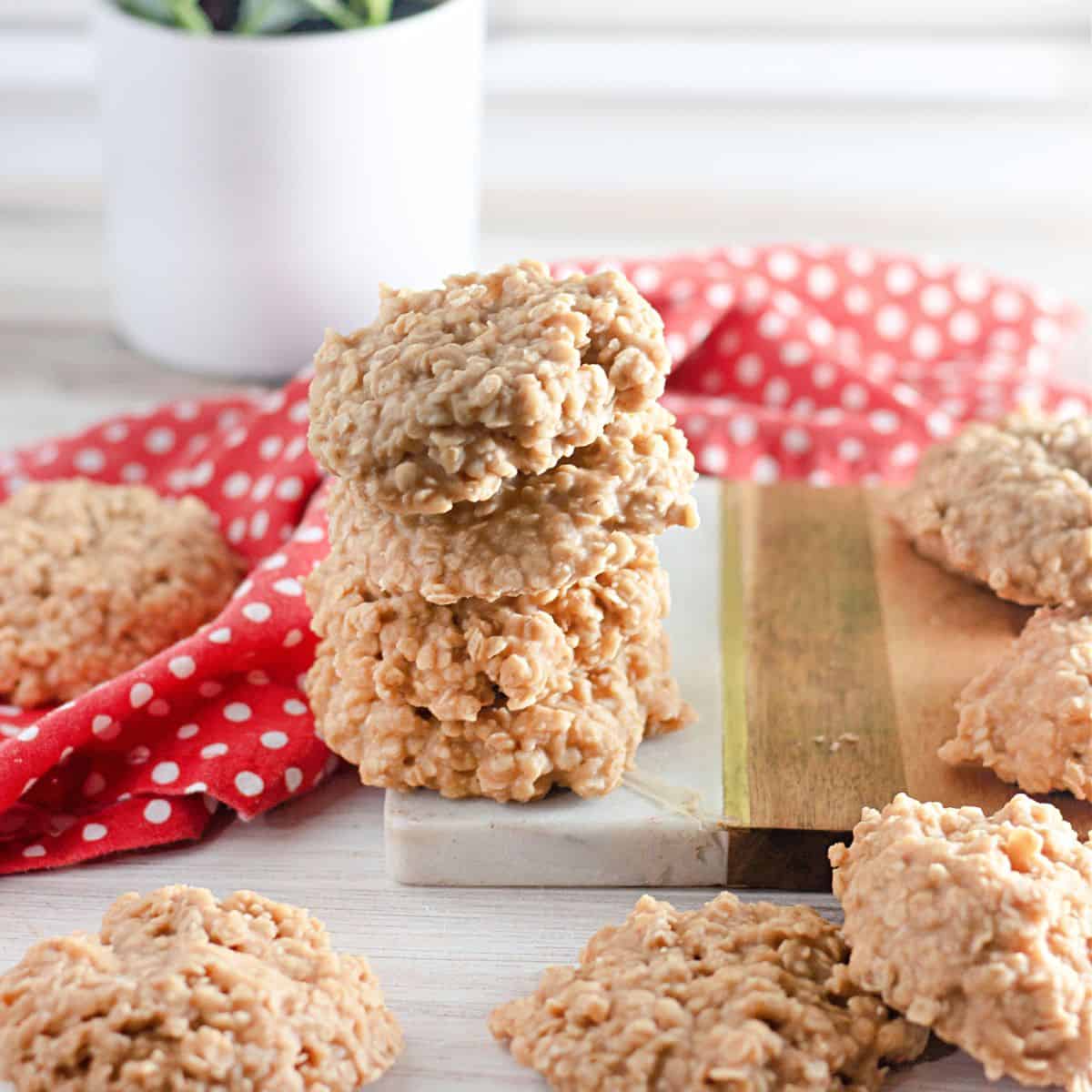 A stack of No Bake Peanut Butter Oatmeal Cookies sits on a marble and wood board, with more cookies and a red polka dot cloth nearby.