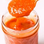 A spoon holding a portion of peach BBQ sauce is placed above a glass jar filled with the vibrant orange-red sauce.