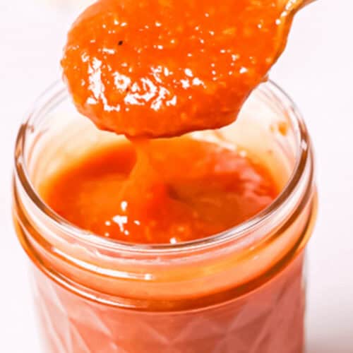 A spoon holding a portion of peach BBQ sauce is placed above a glass jar filled with the vibrant orange-red sauce.
