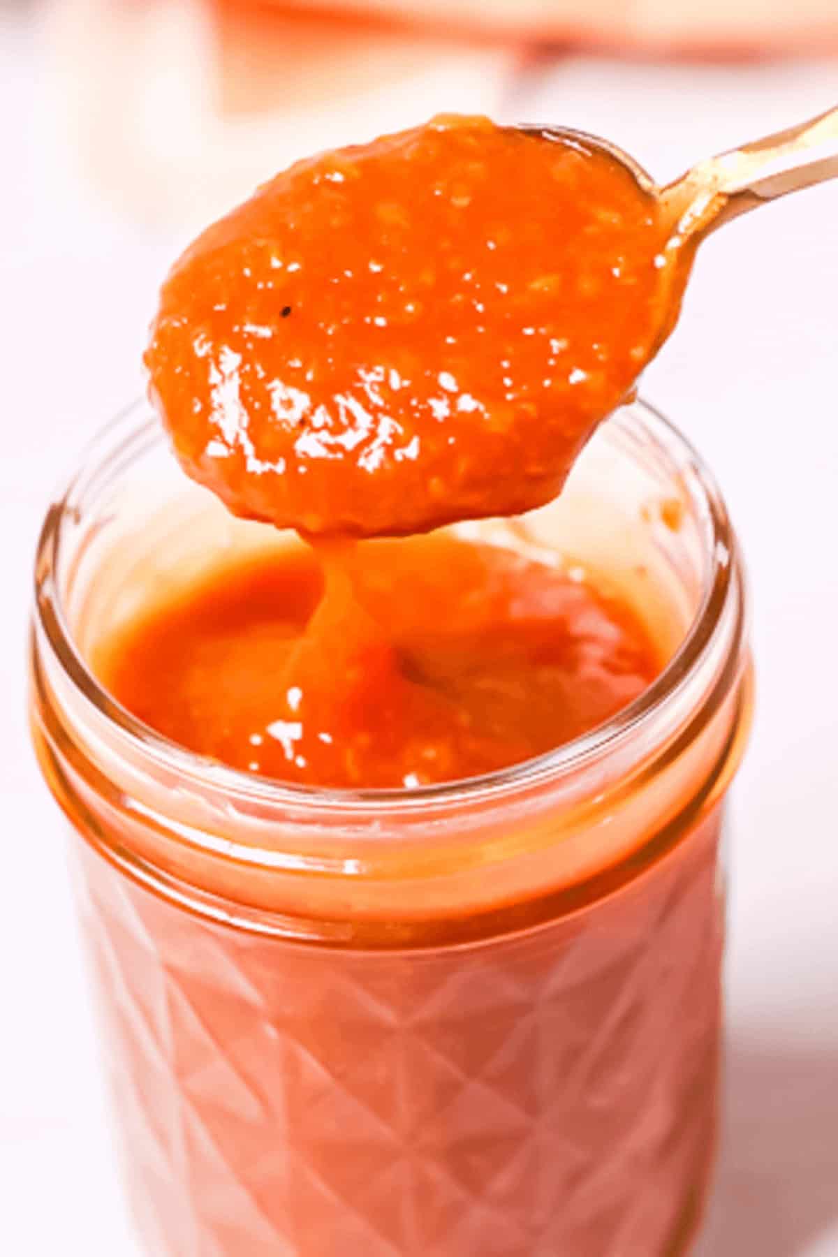 A spoon holding a portion of peach BBQ sauce is placed above a glass jar filled with the vibrant orange-red sauce.