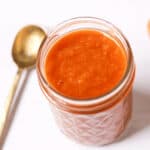 A small glass jar filled with peach BBQ sauce sits on a white surface next to a gold spoon, showcasing its enticing red-orange hue.