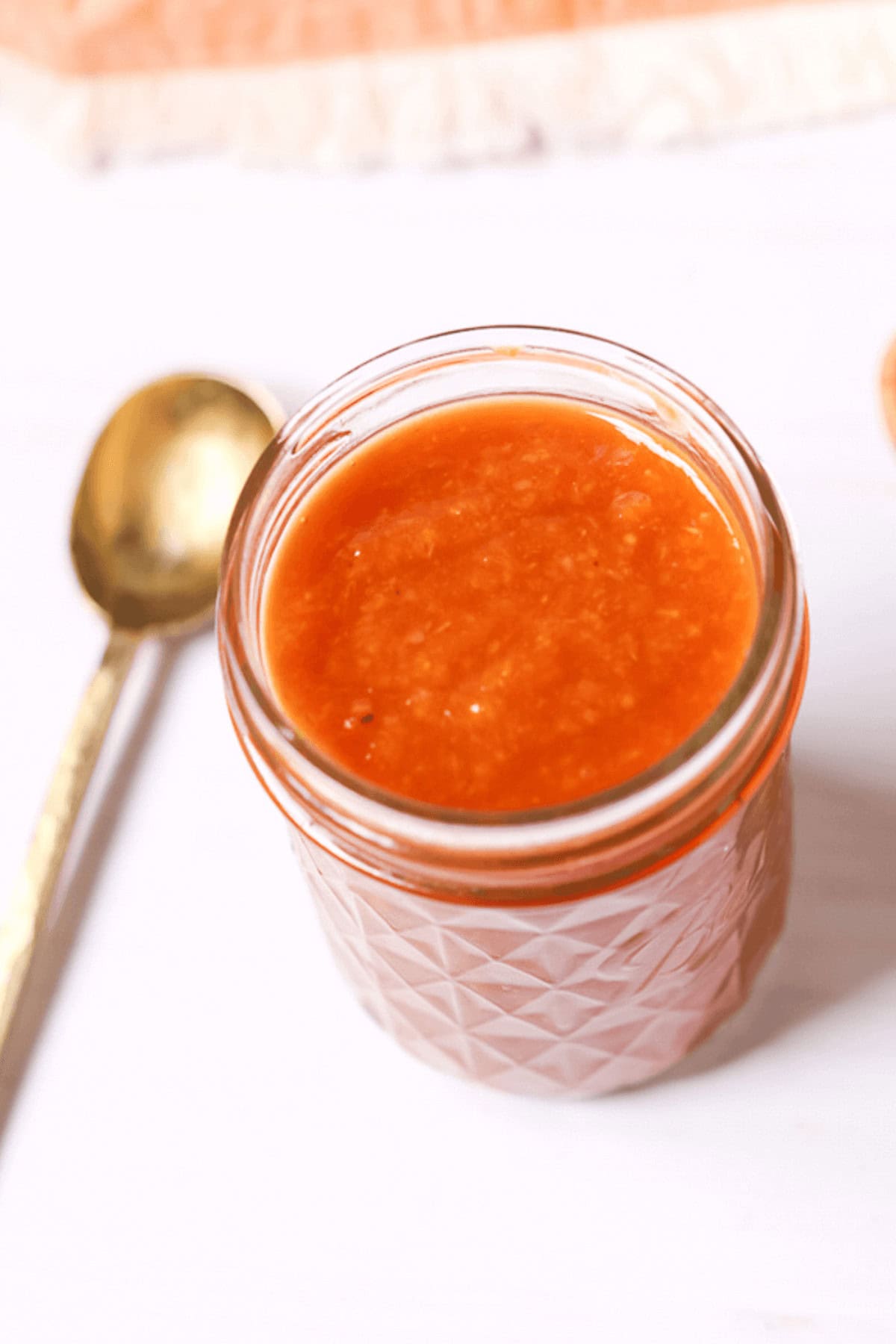 A small glass jar filled with peach BBQ sauce sits on a white surface next to a gold spoon, showcasing its enticing red-orange hue.