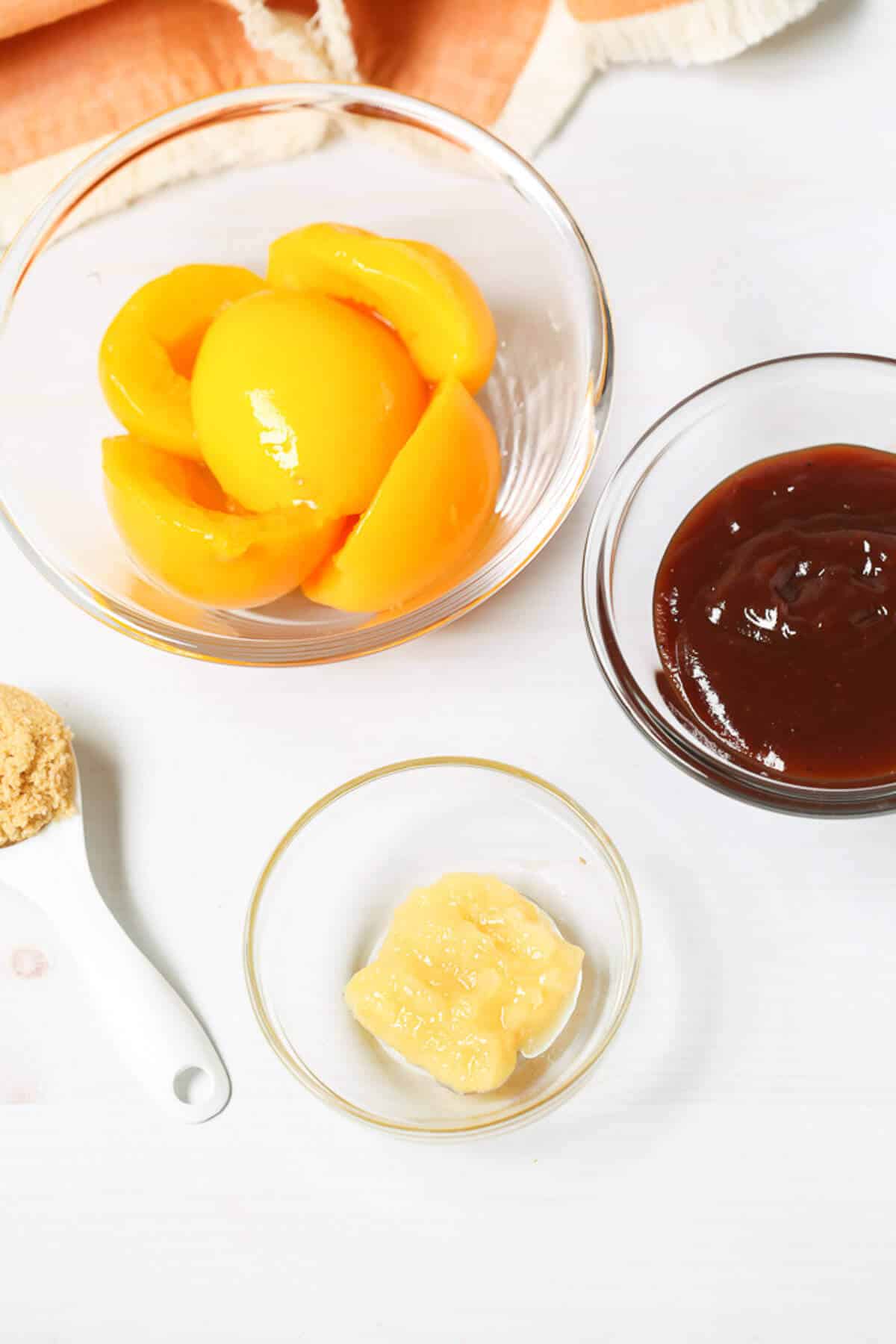 A clear bowl of peach slices, a clear bowl of Peach BBQ Sauce, a clear bowl of minced garlic, and a spoonful of brown sugar are arranged on a white surface.