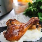 Two BBQ Slow Cooker Drumsticks, perfectly glazed, served on a bed of mashed potatoes with parsley and a glass jar in the background.
