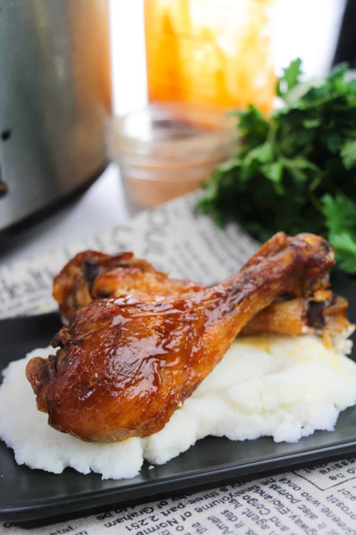 Two BBQ Slow Cooker Drumsticks, perfectly glazed, served on a bed of mashed potatoes with parsley and a glass jar in the background.