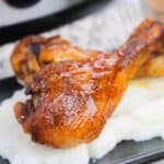 Two BBQ Slow Cooker Drumsticks, glazed to perfection, are placed on a serving of mashed potatoes on a dark plate.