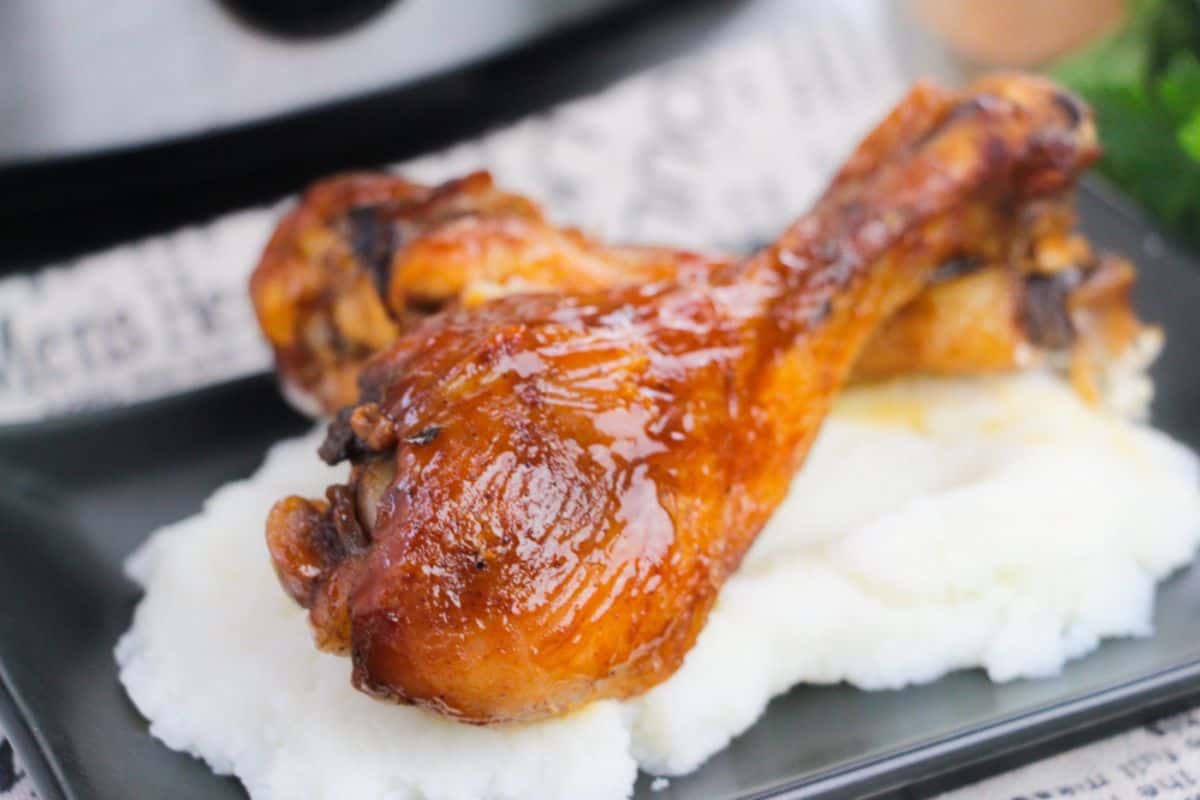 Two BBQ Slow Cooker Drumsticks, glazed to perfection, are placed on a serving of mashed potatoes on a dark plate.
