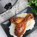 Two cooked BBQ Slow Cooker Drumsticks atop mashed potatoes on a black plate, next to a slow cooker, seasoning jar, fresh parsley, and newspaper print.