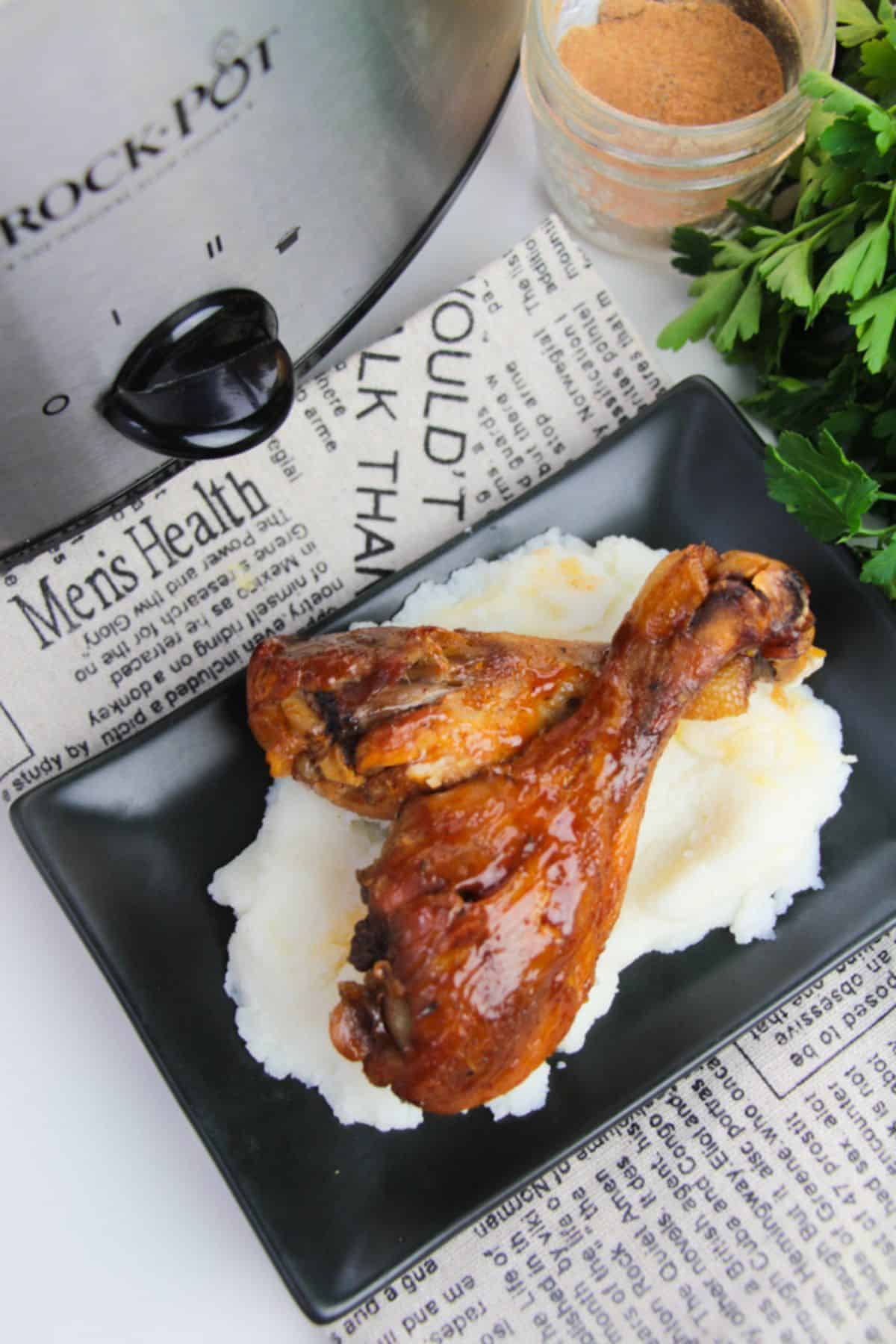 Two cooked BBQ Slow Cooker Drumsticks atop mashed potatoes on a black plate, next to a slow cooker, seasoning jar, fresh parsley, and newspaper print.