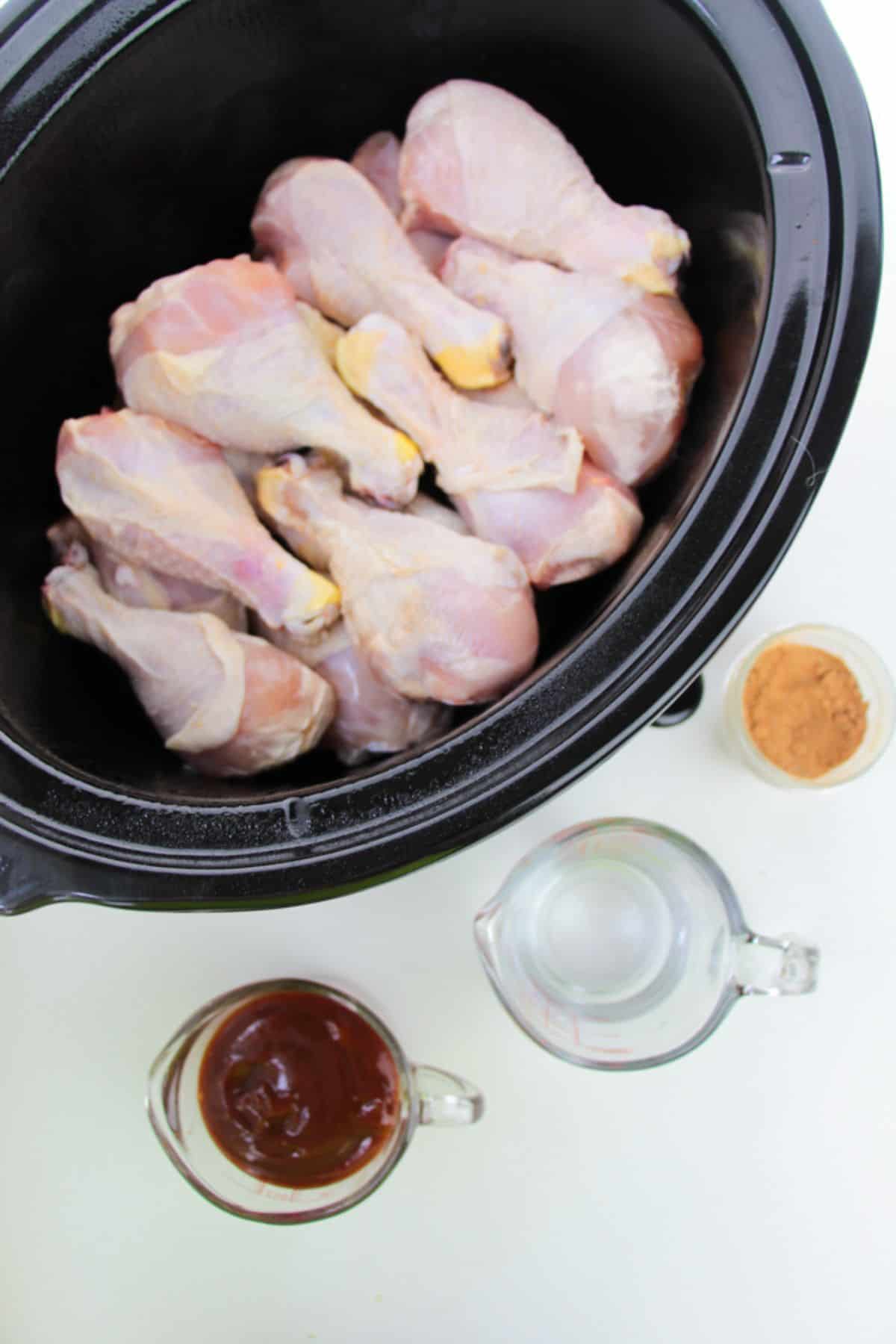 A crock pot with small containers of seasoning, barbecue sauce, and water arranged nearby on a white surface.
