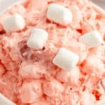 A bowl of pink ambrosia salad, reminiscent of strawberry fluff salad, is topped with mini marshmallows. The creamy texture complements the delightful mix of fruit pieces throughout.