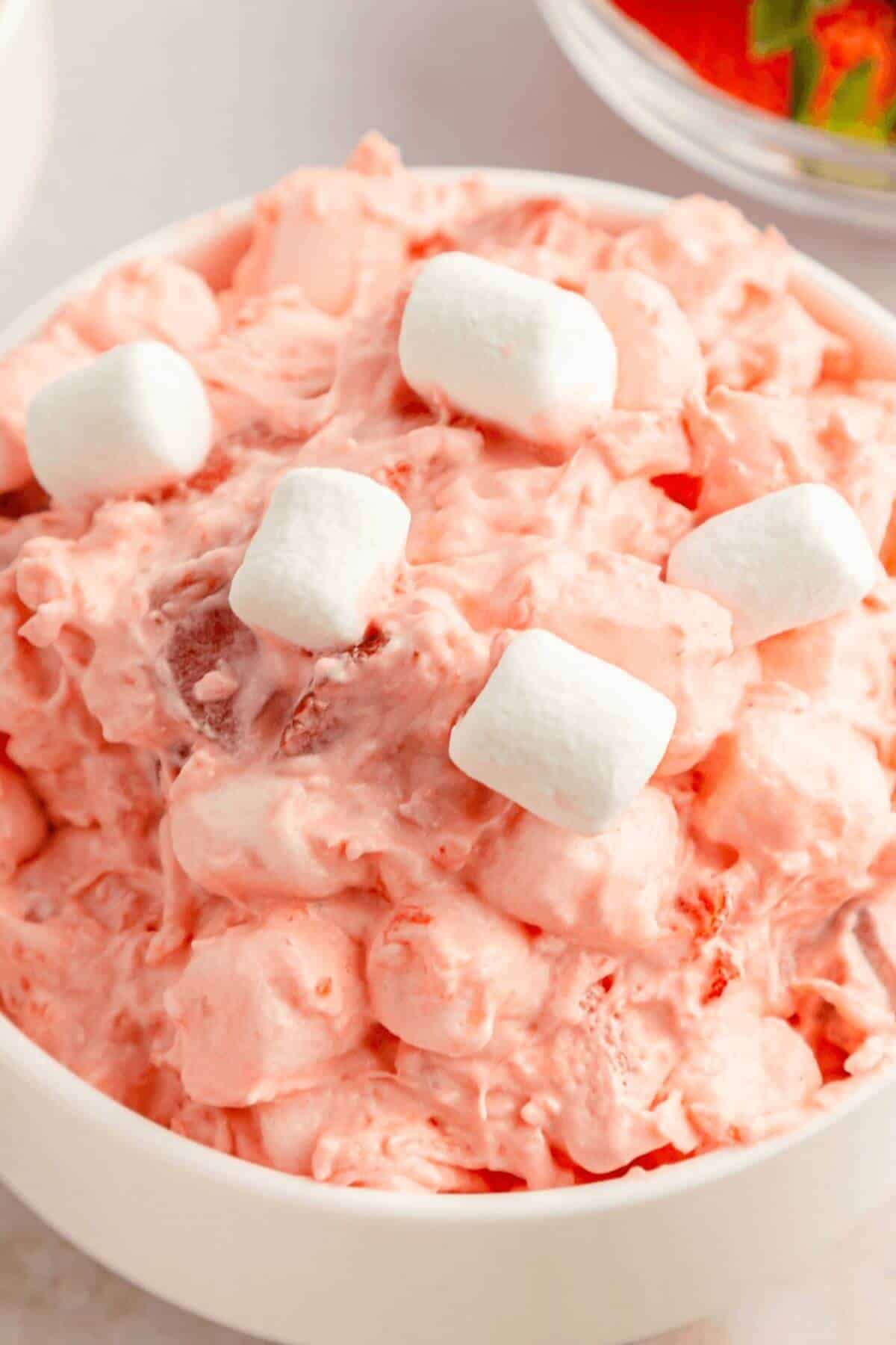 A bowl of pink ambrosia salad, reminiscent of strawberry fluff salad, is topped with mini marshmallows. The creamy texture complements the delightful mix of fruit pieces throughout.
