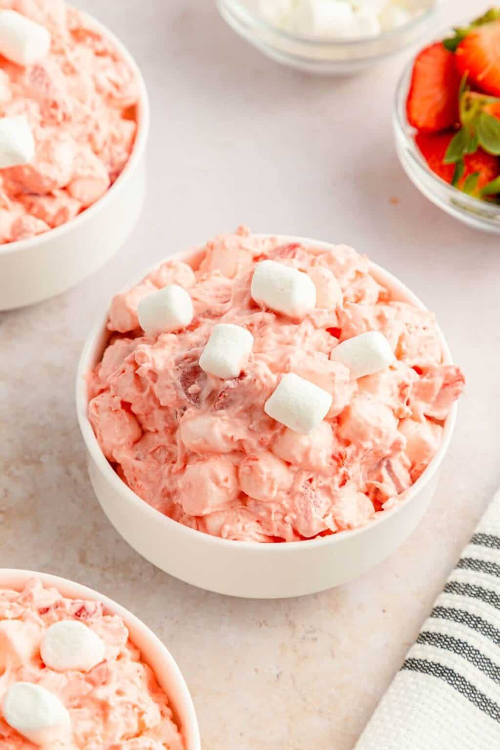 A bowl of pink Strawberry Fluff Salad is topped with marshmallows, while small bowls of juicy strawberries and extra fluff salad sit invitingly in the background.