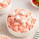 A bowl of pink Strawberry Fluff Salad is topped with marshmallows, while small bowls of juicy strawberries and extra fluff salad sit invitingly in the background.