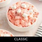 A bowl of Strawberry Fluff Salad topped with mini marshmallows sits next to another bowl and a small dish of strawberries, all above a black stripe labeled "Classic Strawberry Fluff Salad.