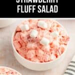 A delightful bowl of Strawberry Fluff Salad, brimming with pink hues and crowned with marshmallows, rests on a light surface beside an elegant striped cloth.