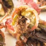 Close-up of a stuffed pepper wrapped in bacon, showcasing jalapeño, cream cheese, and mushrooms, all secured by a toothpick. Similar mouthwatering appetizers are visible in the background.