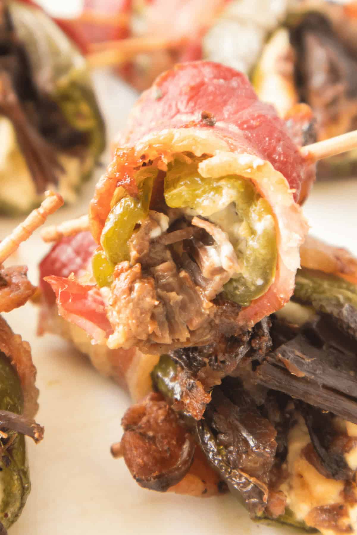 Close-up of a stuffed pepper wrapped in bacon, showcasing jalapeño, cream cheese, and mushrooms, all secured by a toothpick. Similar mouthwatering appetizers are visible in the background.