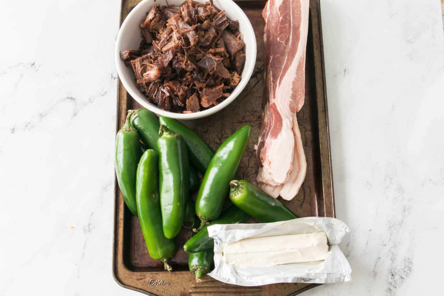 A baking tray showcases a bowl of shredded meat, raw bacon slices, jalapeños, and a wrapped block of cream cheese on a marble surface—perfect ingredients ready to create Texas Twinkies.