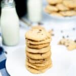 A stack of bakery style chocolate chip cookies on a white plate, with bottles of milk and more cookies on a stand in the background.