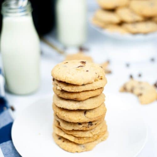 A stack of bakery style chocolate chip cookies on a white plate, with bottles of milk and more cookies on a stand in the background.
