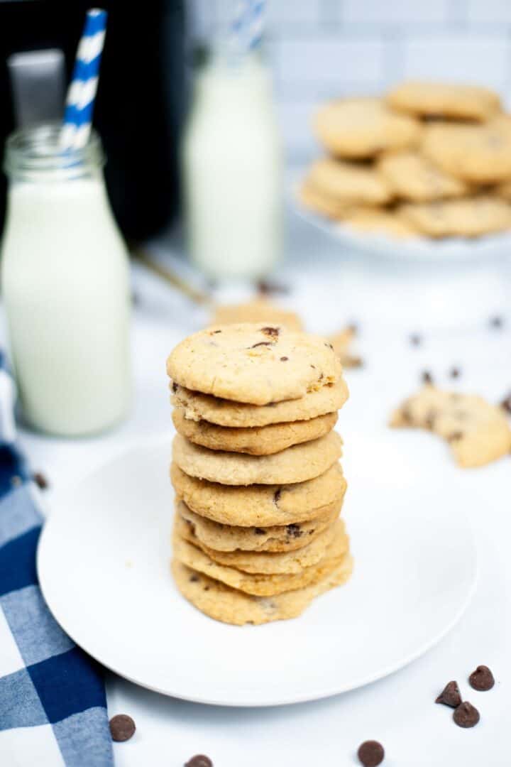 A stack of bakery style chocolate chip cookies on a white plate, with bottles of milk and more cookies on a stand in the background.