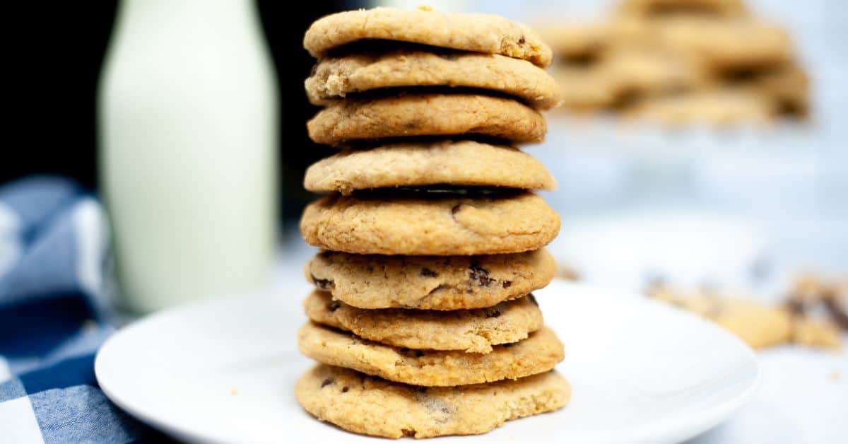A stack of bakery style chocolate chip cookies sits on a white plate with a bottle of milk and a blue checkered cloth in the background.