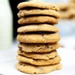 A stack of bakery style chocolate chip cookies sits on a white plate with a bottle of milk and a blue plaid cloth in the background.