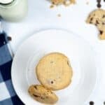 A plate with two bakery style chocolate chip cookies, surrounded by a glass bottle of milk with straws, scattered brown sugar, chocolate chips, and a blue checkered napkin.