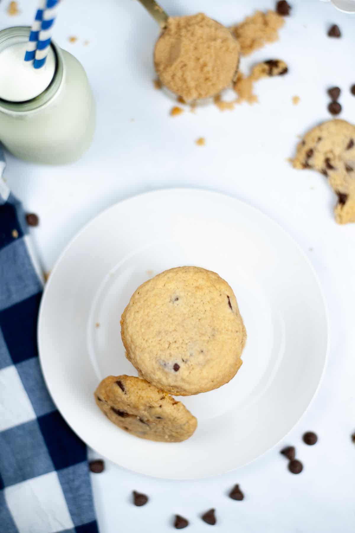 A plate with two bakery style chocolate chip cookies, surrounded by a glass bottle of milk with straws, scattered brown sugar, chocolate chips, and a blue checkered napkin.