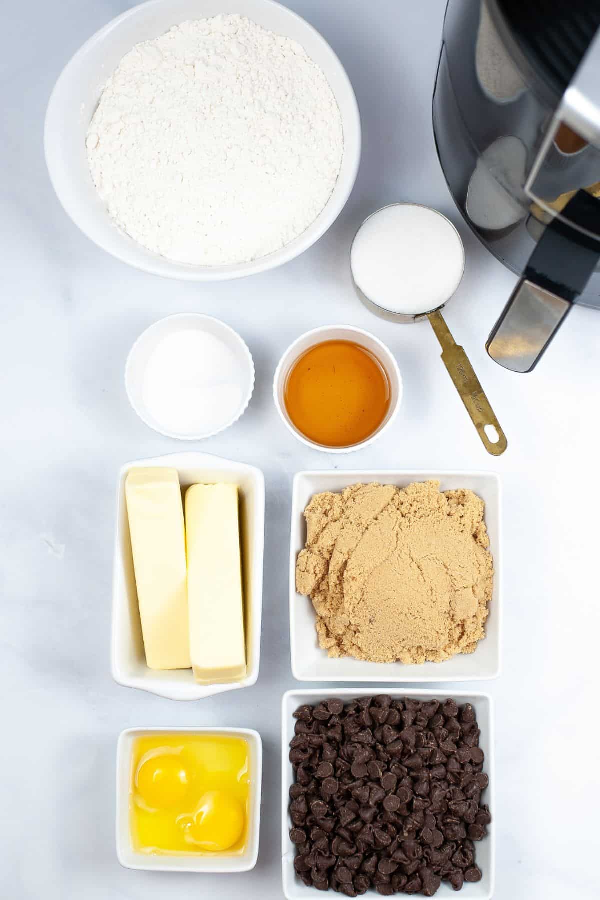Baking ingredients arranged on a white surface: flour, sugar, salt, honey, butter, brown sugar, eggs, chocolate chips.