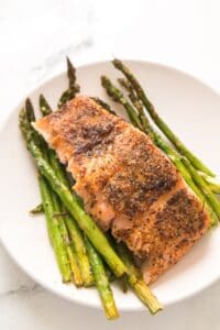 A delicious piece of seasoned, cooked salmon is served atop roasted asparagus spears on a white plate, creating a classic Salmon and Asparagus pairing that’s both elegant and flavorful.