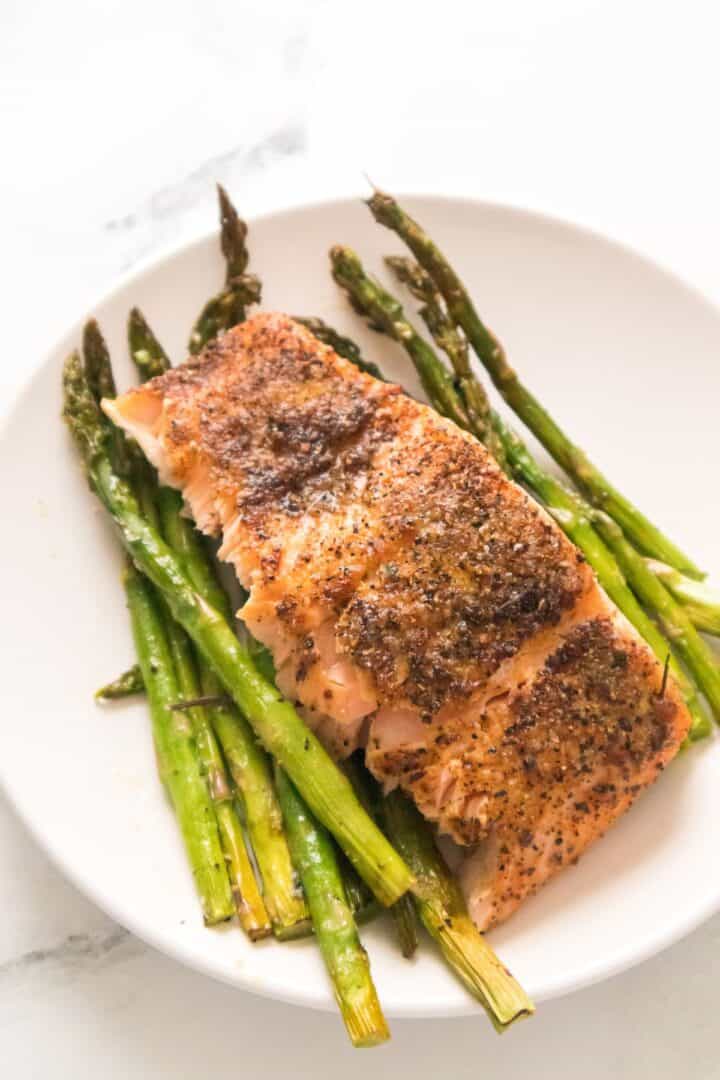 A delicious piece of seasoned, cooked salmon is served atop roasted asparagus spears on a white plate, creating a classic Salmon and Asparagus pairing that’s both elegant and flavorful.