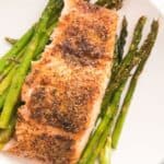 A cooked salmon fillet seasoned with spices is served on a white plate, beautifully paired with roasted asparagus spears arranged underneath, creating a classic Salmon and Asparagus dish.