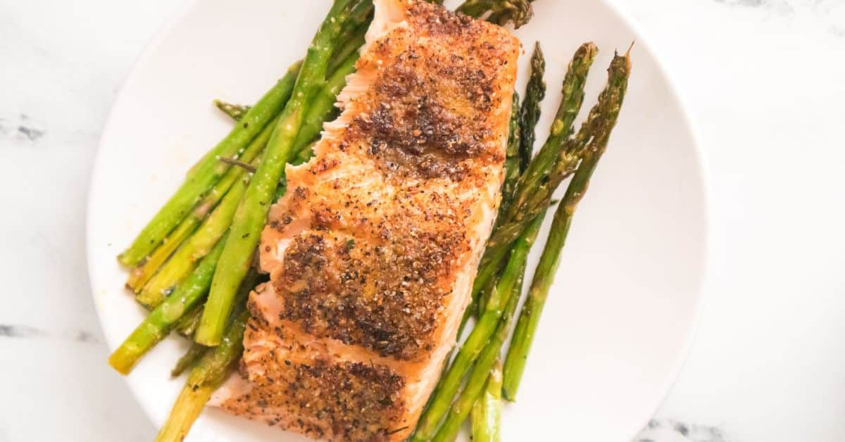 A cooked salmon fillet seasoned with spices is served on a white plate, beautifully paired with roasted asparagus spears arranged underneath, creating a classic Salmon and Asparagus dish.