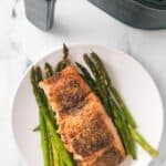 A plate with a cooked fish and featuring tender roasted vegetables sits next to an air fryer on a white surface.
